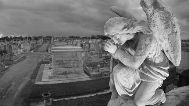 A praying statue kneels on top of a tomb in New Orleans under moody skies. High point of view to contain the cemetery landscape in the background. credit: istock one time use for Traveller only